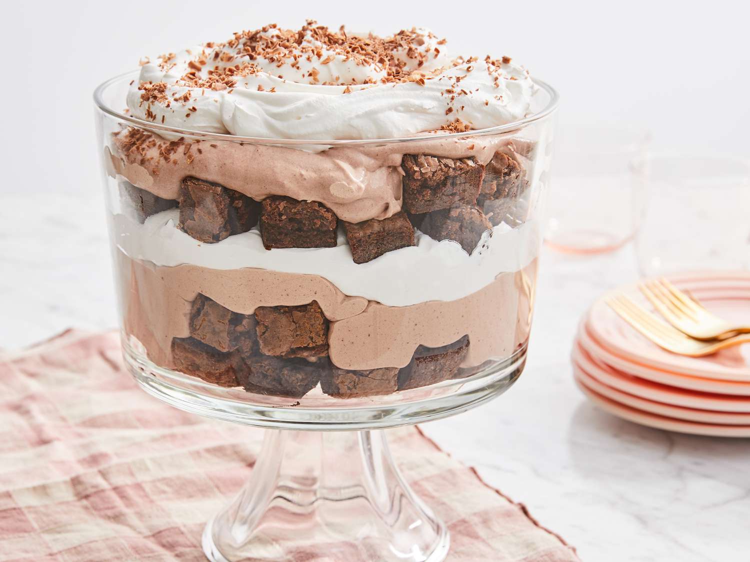 Ingredients for chocolate trifle arranged on a kitchen counter including brownie chunks, milk, eggs, chopped chocolate, cocoa powder, heavy cream, raspberries, and sugar