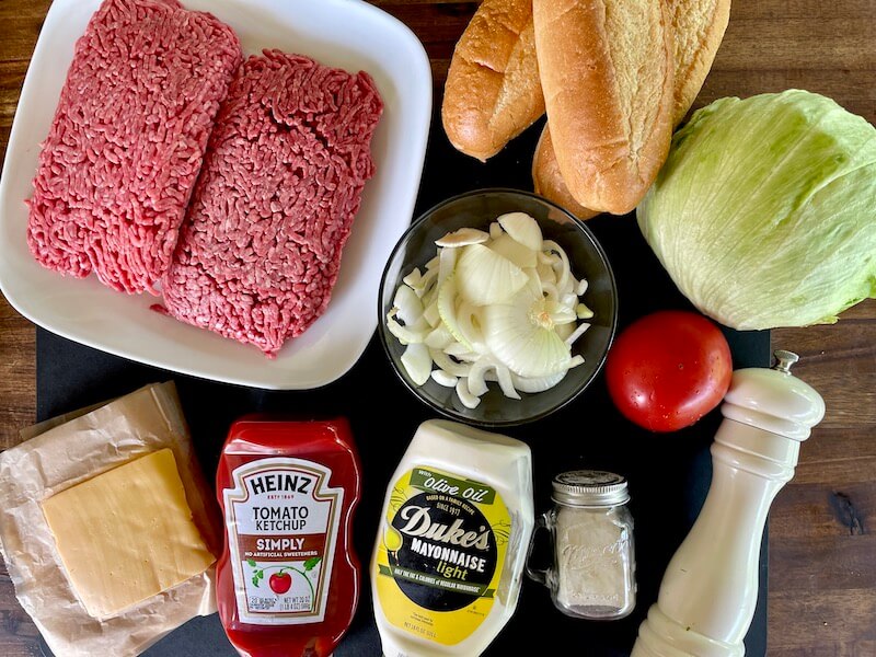 Ingredients for chopped cheese laid out on a kitchen counter: ground beef, sliced onion, American cheese, hero rolls, lettuce, tomato, mayonnaise, ketchup, and seasonings