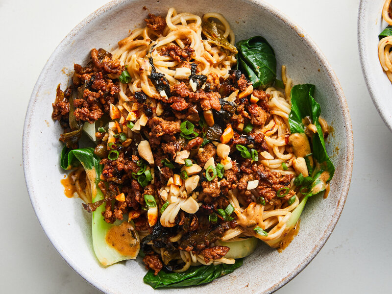 Ingredients for dan dan mian arranged on a kitchen counter including noodles, ground pork, tahini, soy sauce, chili oil, garlic, ginger, scallions, bok choy, and peanuts