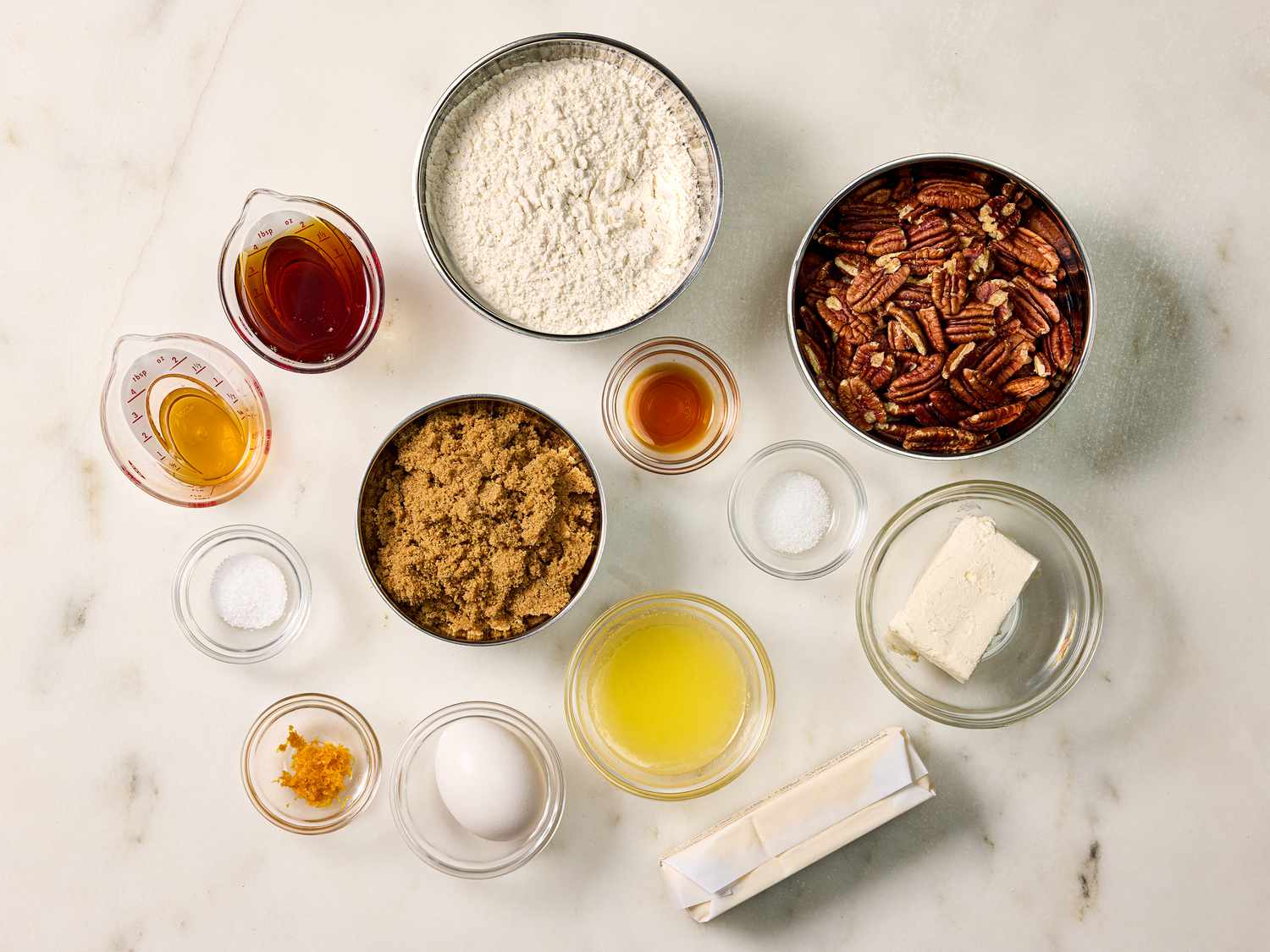 Ingredients for pecan tassies laid out on a tidy kitchen counter including cream cheese, butter, brown sugar, eggs, vanilla, pecans, and a mini muffin pan
