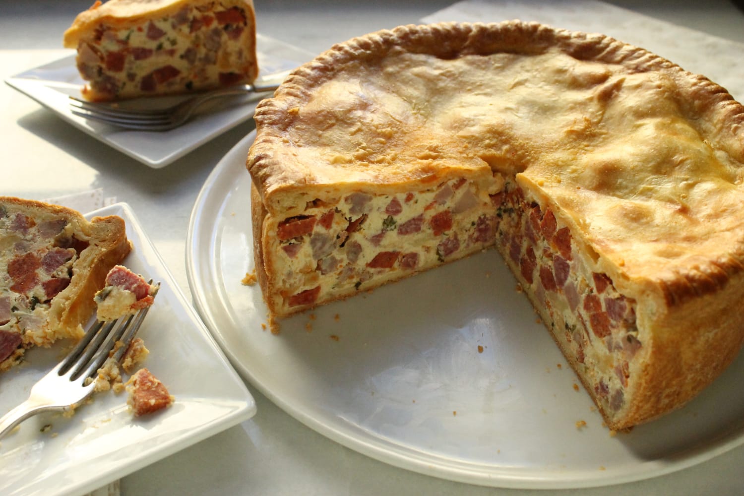 Ingredients for pizza rustica arranged on a light stone kitchen counter: ricotta, eggs, salami, ham, mozzarella, Pecorino Romano, parsley, flour, butter, and a lemon