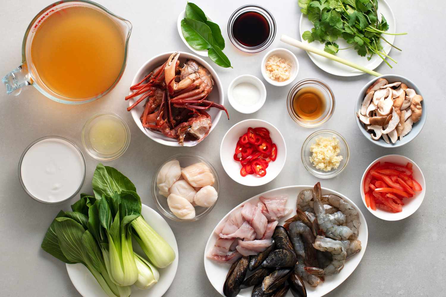 Ingredients for seafood soup arranged on a kitchen counter, including shrimp, cod, clams, fennel, canned tomatoes, garlic, herbs, lemon, and stock