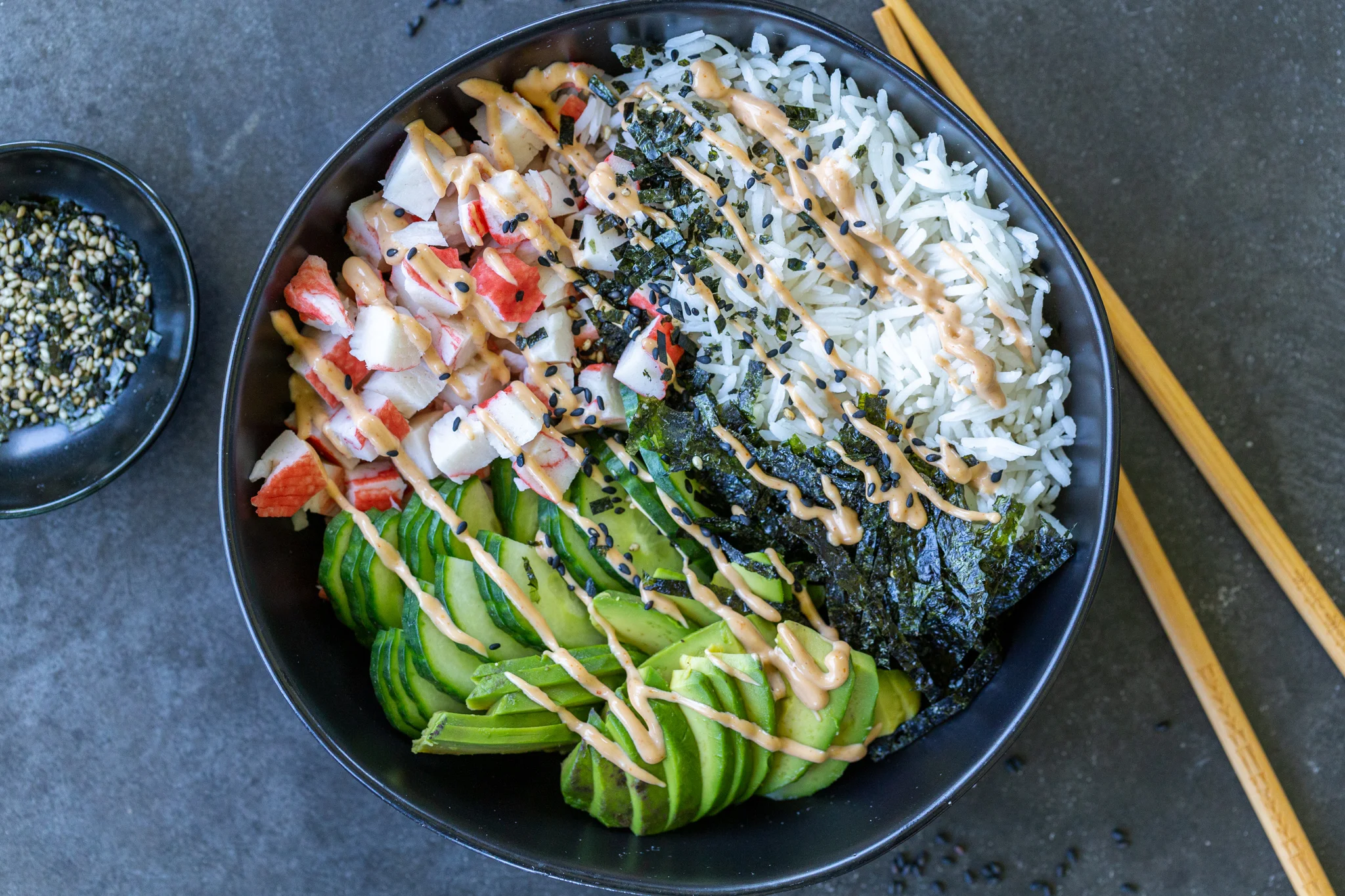 Ingredients for sushi bowls arranged on a kitchen counter, including salmon, cooked rice, cucumbers, avocado, nori sheets, rice vinegar, soy sauce, mayonnaise, sriracha, and sesame seeds