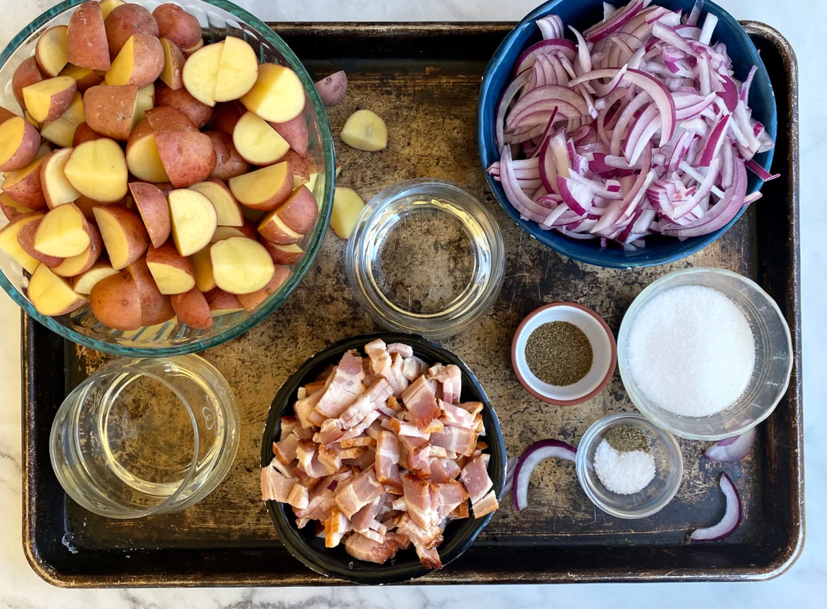 Ingredients for warm German potato salad on a kitchen counter, including small Yukon Gold or red potatoes, bacon, onion, Dijon mustard, apple cider vinegar, sugar, parsley, and salt and pepper