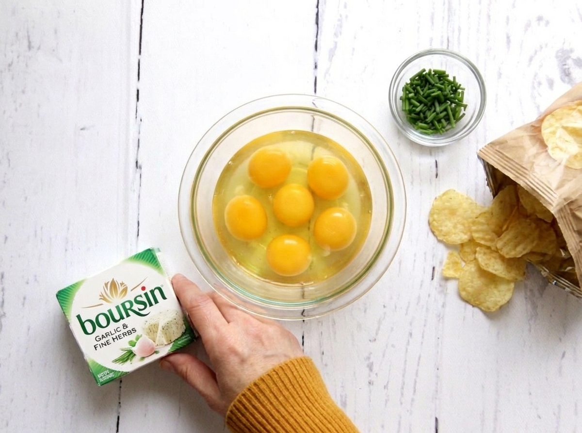 Ingredients on a kitchen counter: eggs in a bowl, a package of Boursin garlic and fine herbs, a small bowl of chopped chives, and a bag of potato chips