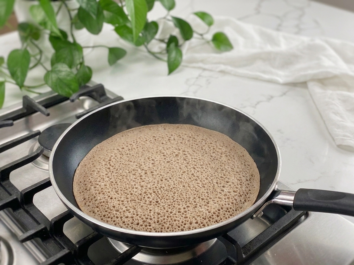Injera batter cooking in a wide nonstick skillet with lots of small bubbles forming across the surface