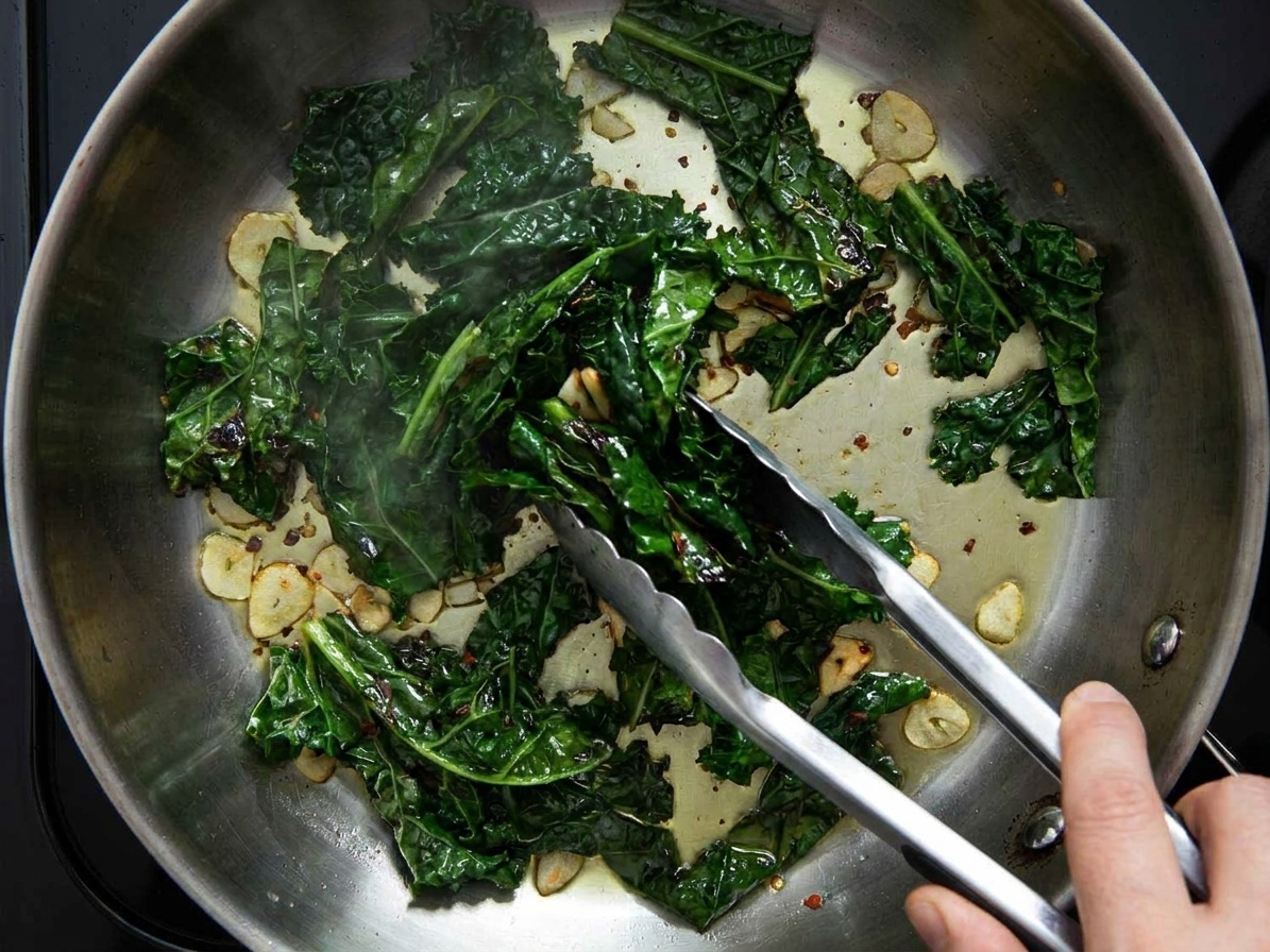 Kale ribbons being tossed with tongs in a hot skillet as they wilt and darken in color
