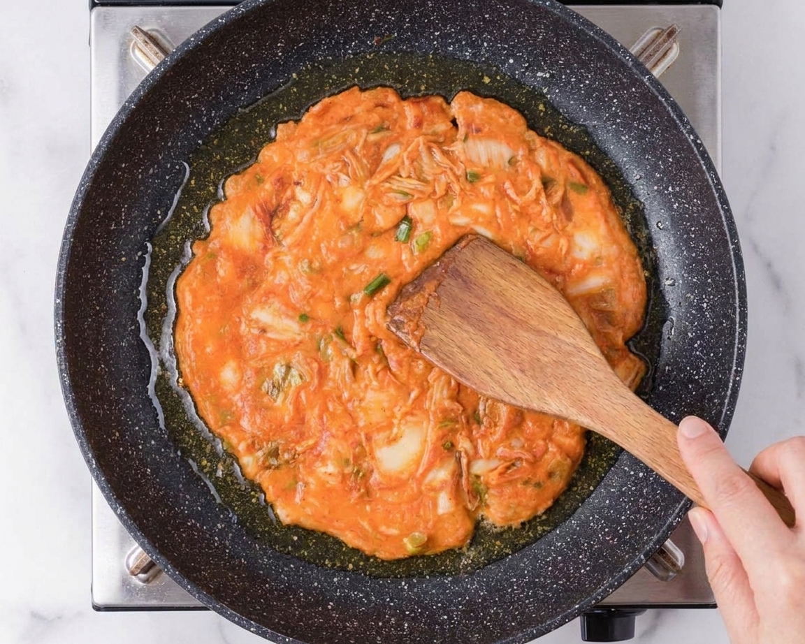 Kimchi pancake batter being spread into a round in a hot oiled skillet, with visible kimchi pieces and scallions