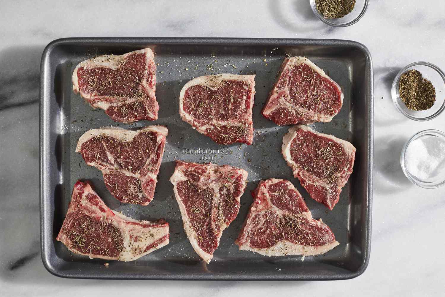 Lamb loin steaks being patted dry and seasoned with salt and black pepper on a cutting board beside a cast iron skillet