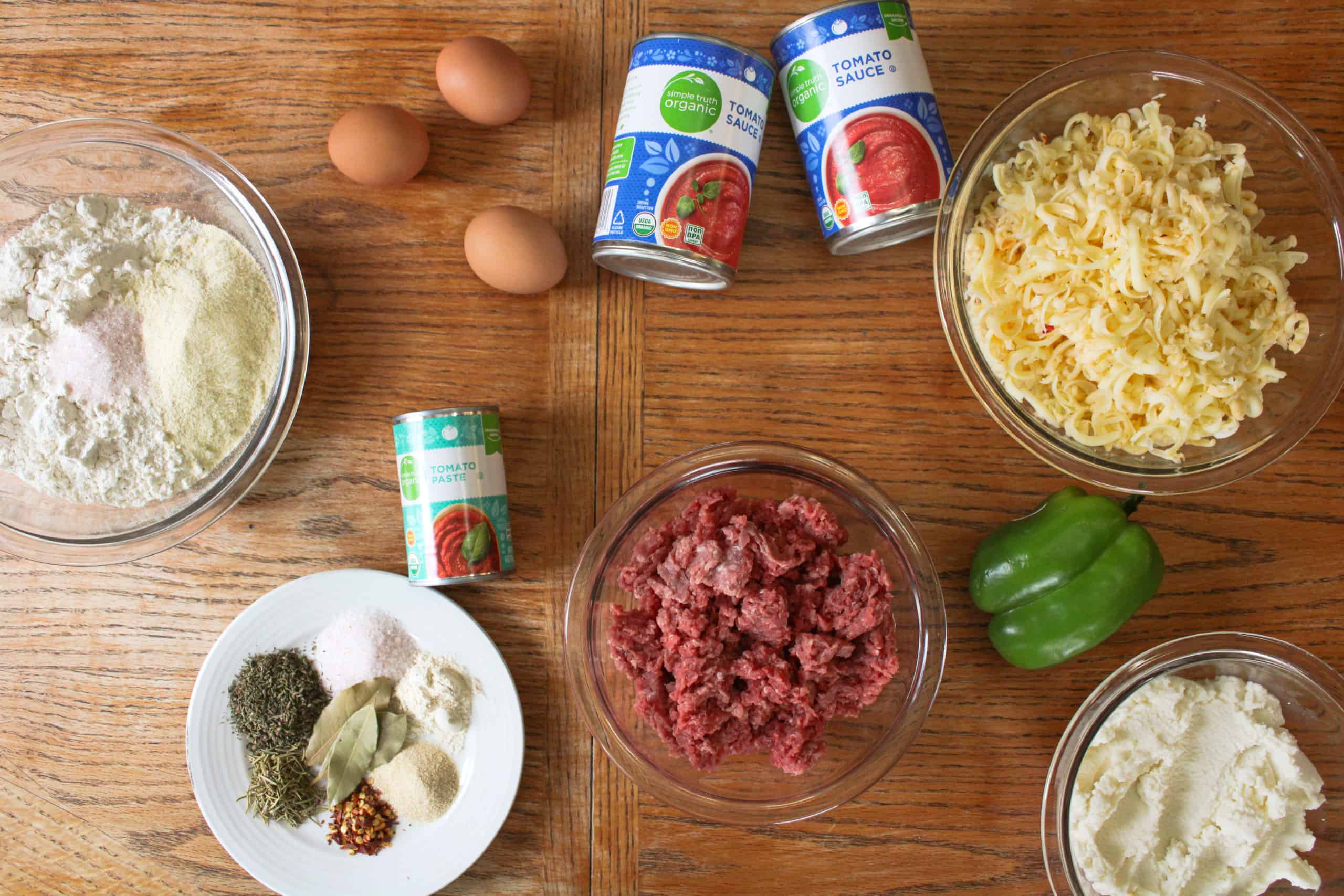 Lasagna ingredients arranged on a home kitchen counter, including ground beef, Italian sausage, canned tomatoes, onions, garlic, milk, butter, flour, lasagna noodles, Parmesan, and mozzarella in warm natural light