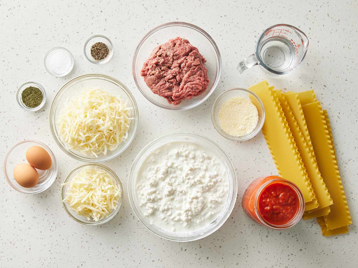 Lasagna ingredients arranged on a kitchen counter including lasagna noodles, ground beef, jarred marinara, ricotta, mozzarella, parmesan, eggs, and herbs