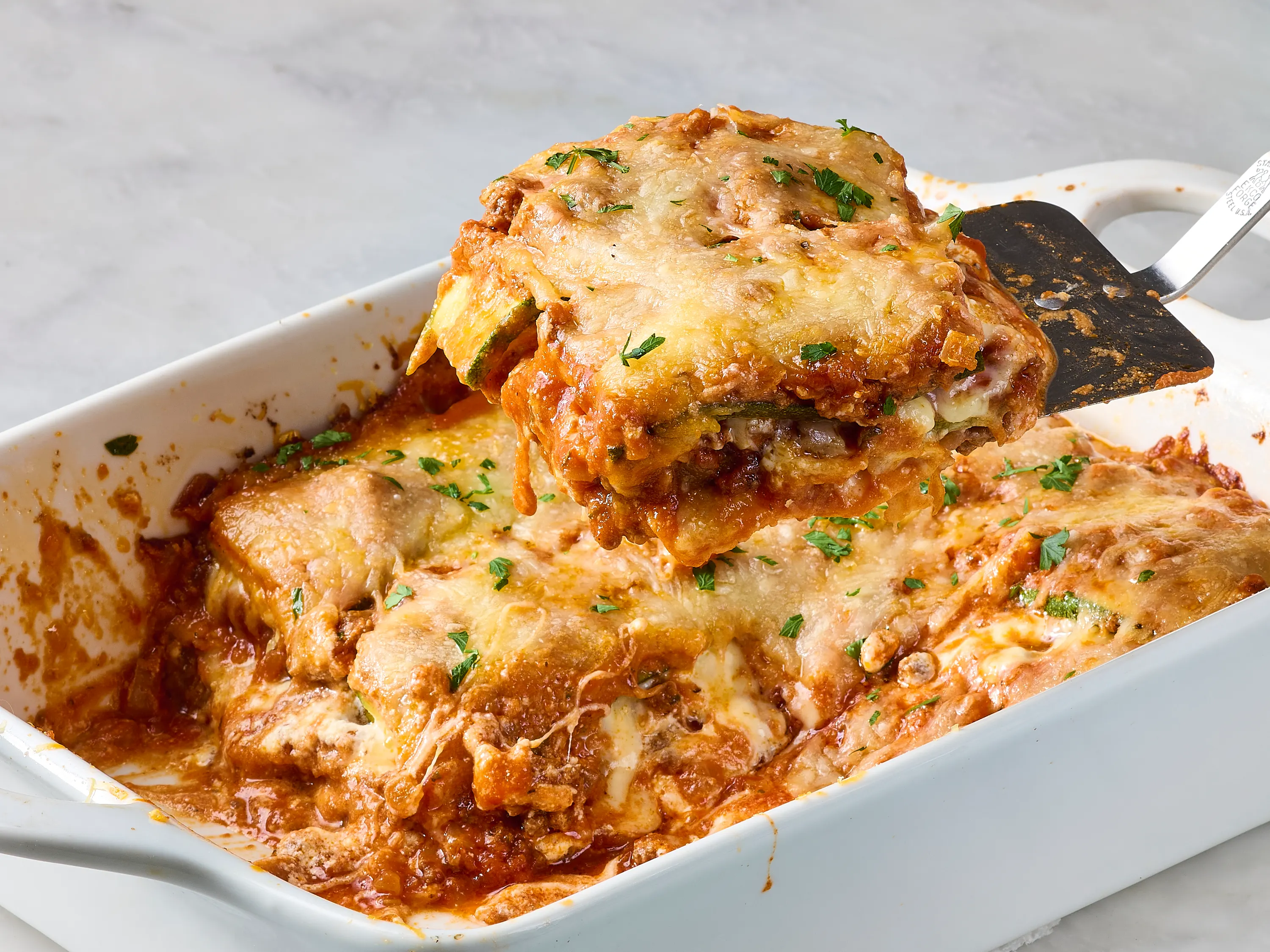 Lasagna ingredients arranged on a kitchen counter including ricotta, mozzarella, marinara sauce, lasagna noodles, zucchini, mushrooms, spinach, and a bowl of grated Parmesan