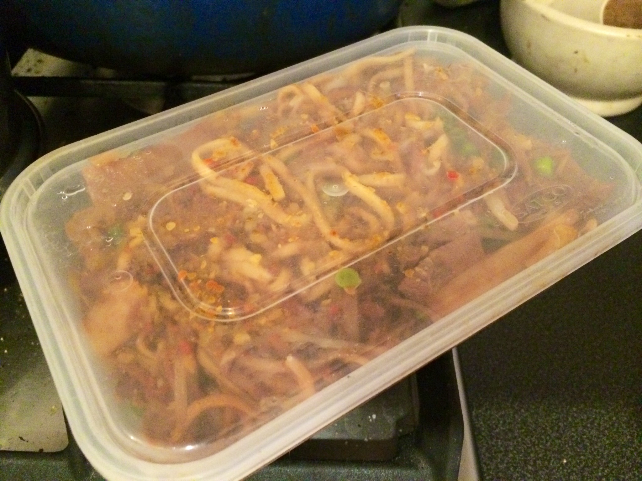 Leftover beef chow fun in a clear airtight container on a kitchen counter beside a small bowl of sauce