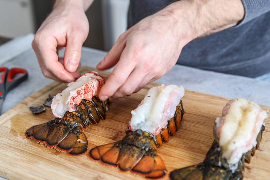 Lobster tail shell being cut lengthwise with kitchen shears on a wooden cutting board under warm kitchen lighting