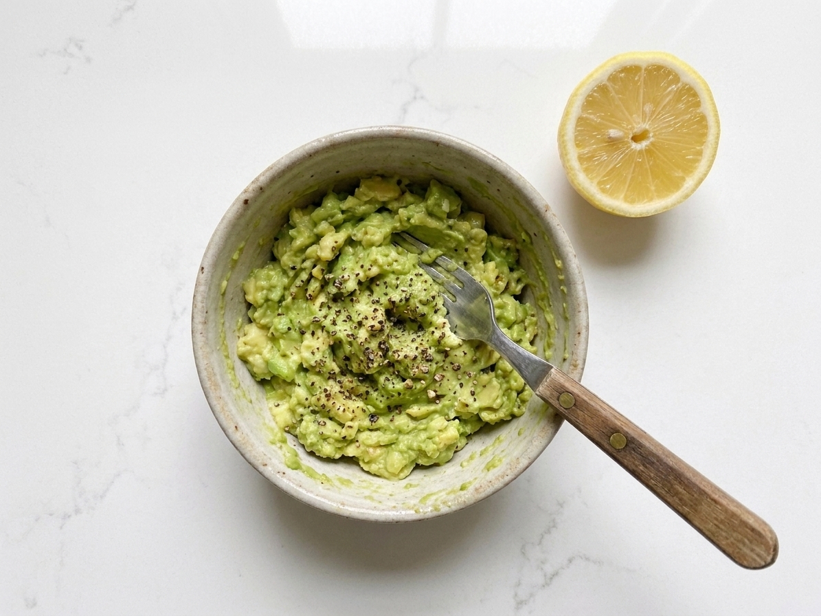 Mashed avocado in a small bowl with a fork, lemon half nearby, and visible black pepper on a kitchen counter