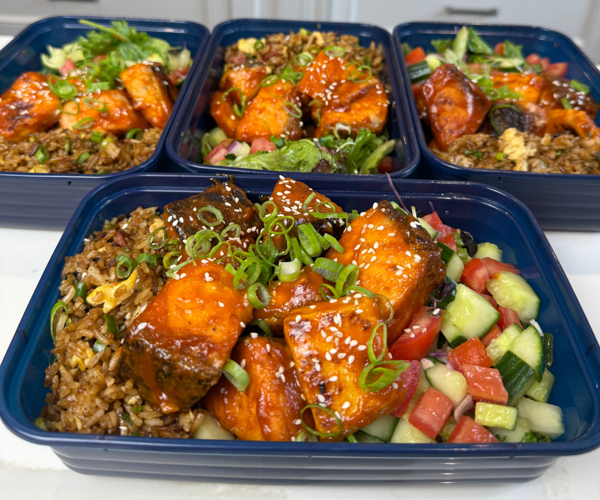 Meal prep containers filled with salmon, broccoli, and rice on a kitchen counter with small containers of lemon yogurt sauce