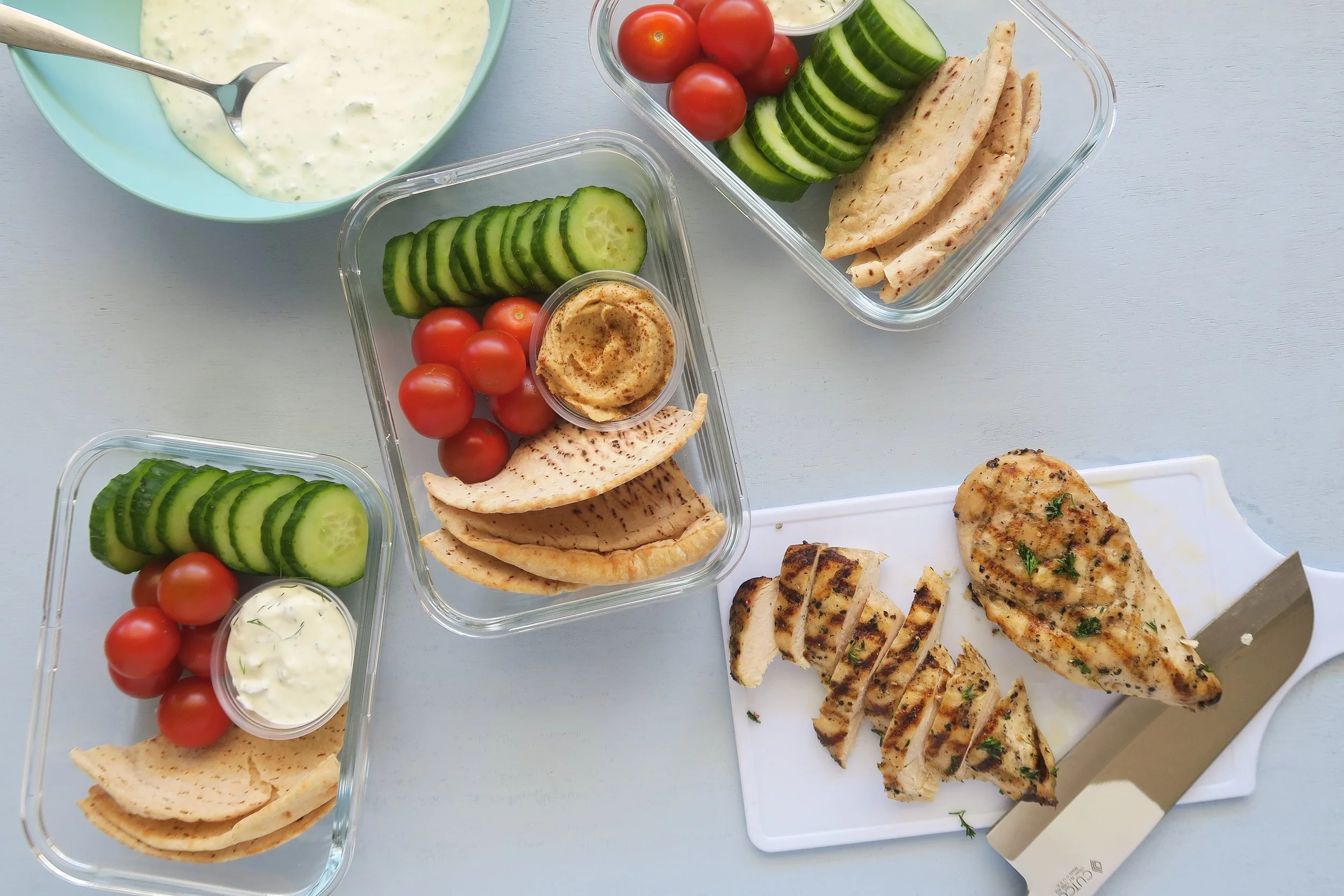 Meal prep containers with sliced chicken souvlaki, chopped tomatoes, cucumbers, and pita wedges arranged on a kitchen counter