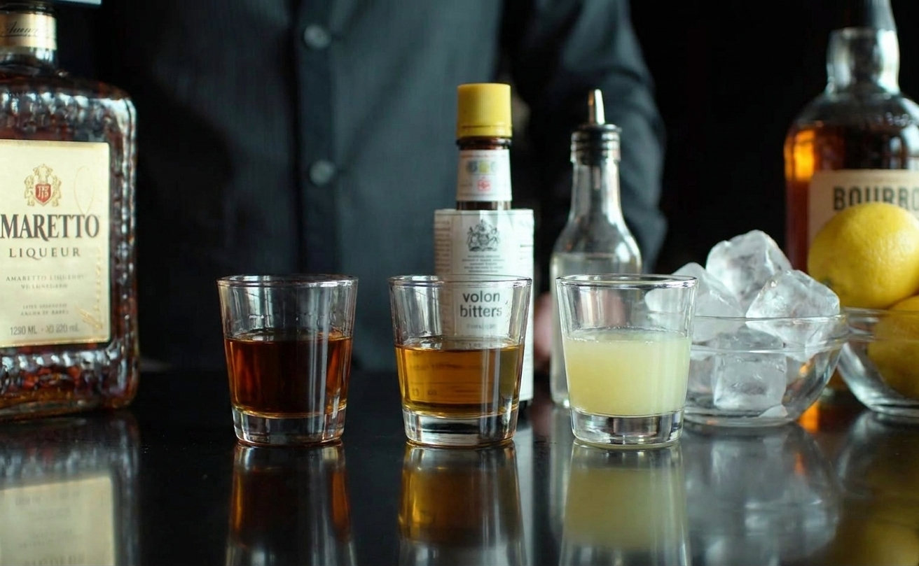 Measured amaretto, bourbon, and fresh lemon juice in small glass cups with bitters and ice nearby on a kitchen counter