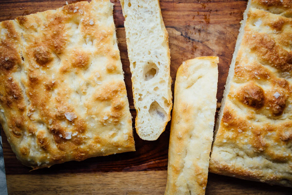 Measured bread flour in a bowl with a small dish of salt and a jar of bubbly sourdough starter on a kitchen counter with warm light
