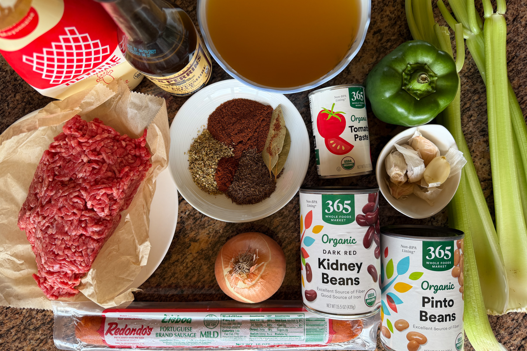Measured chili ingredients including ground beef, canned beans, diced tomatoes, tomato sauce, onions, celery, and spices laid out on a bright kitchen counter