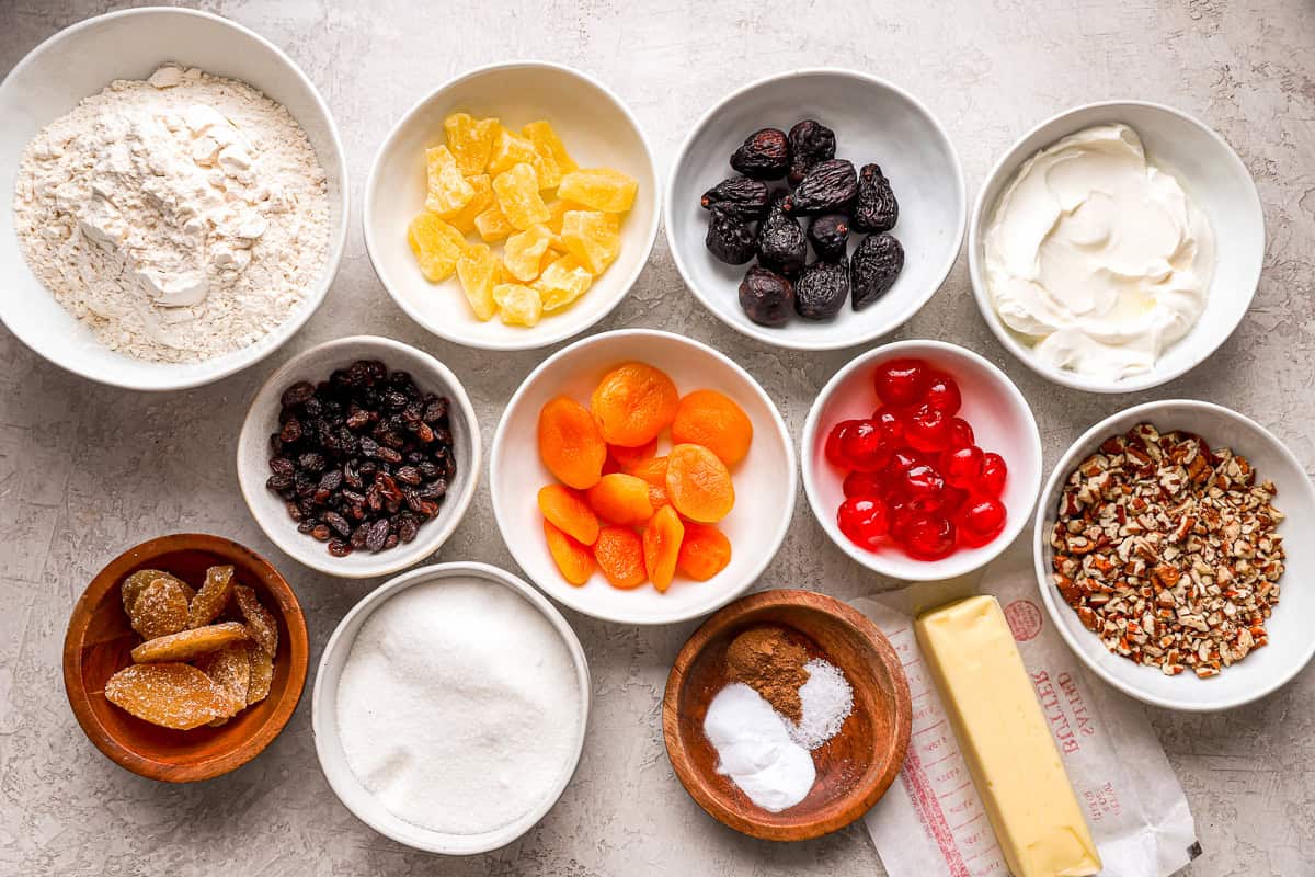 Measured dried fruits, chopped nuts, flour, spices, and citrus zest arranged in small bowls on a clean kitchen counter, ready for baking