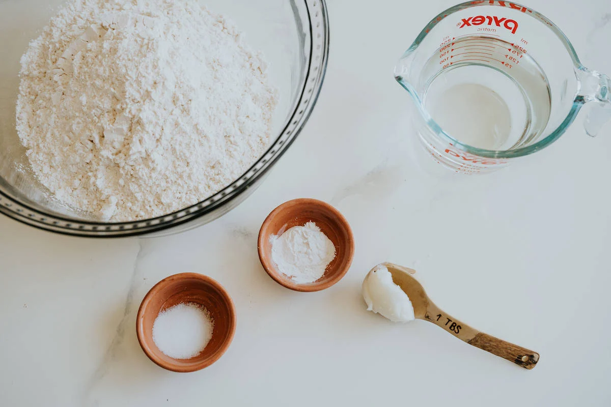 Measured flour, salt, baking powder, and a scoop of lard in small bowls on a tidy kitchen counter with warm natural light