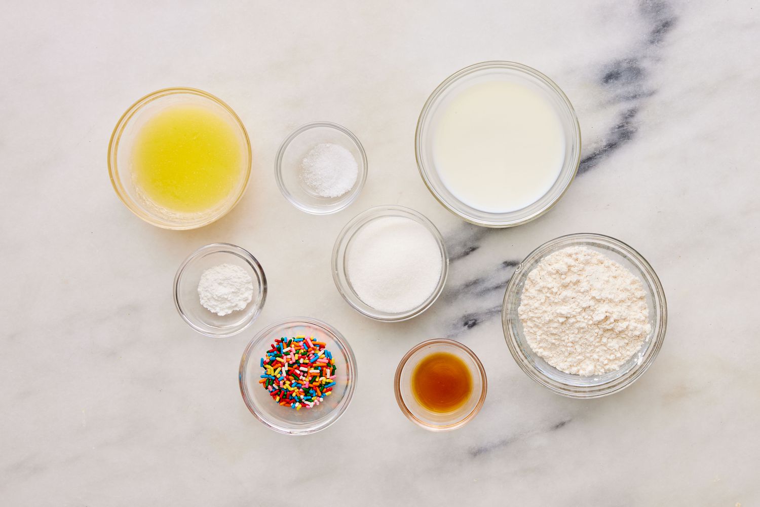 Measured flour, sugar, baking powder, milk, melted butter, vanilla extract, and a cracked egg on a simple kitchen counter beside a large microwave-safe mug