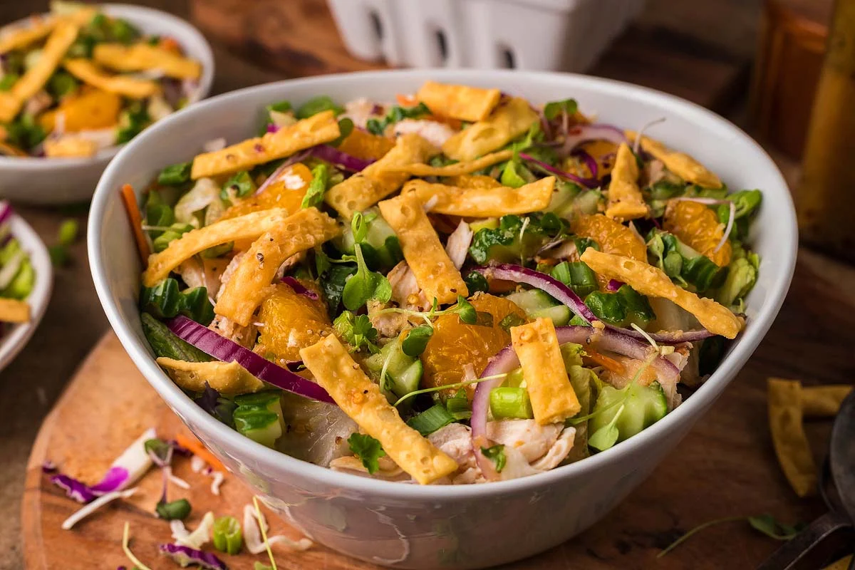 Measured ingredients for Asian chicken salad arranged on a wooden cutting board, including shredded cabbage, carrots, cucumbers, scallions, cilantro, toasted sesame seeds, and cooked chicken