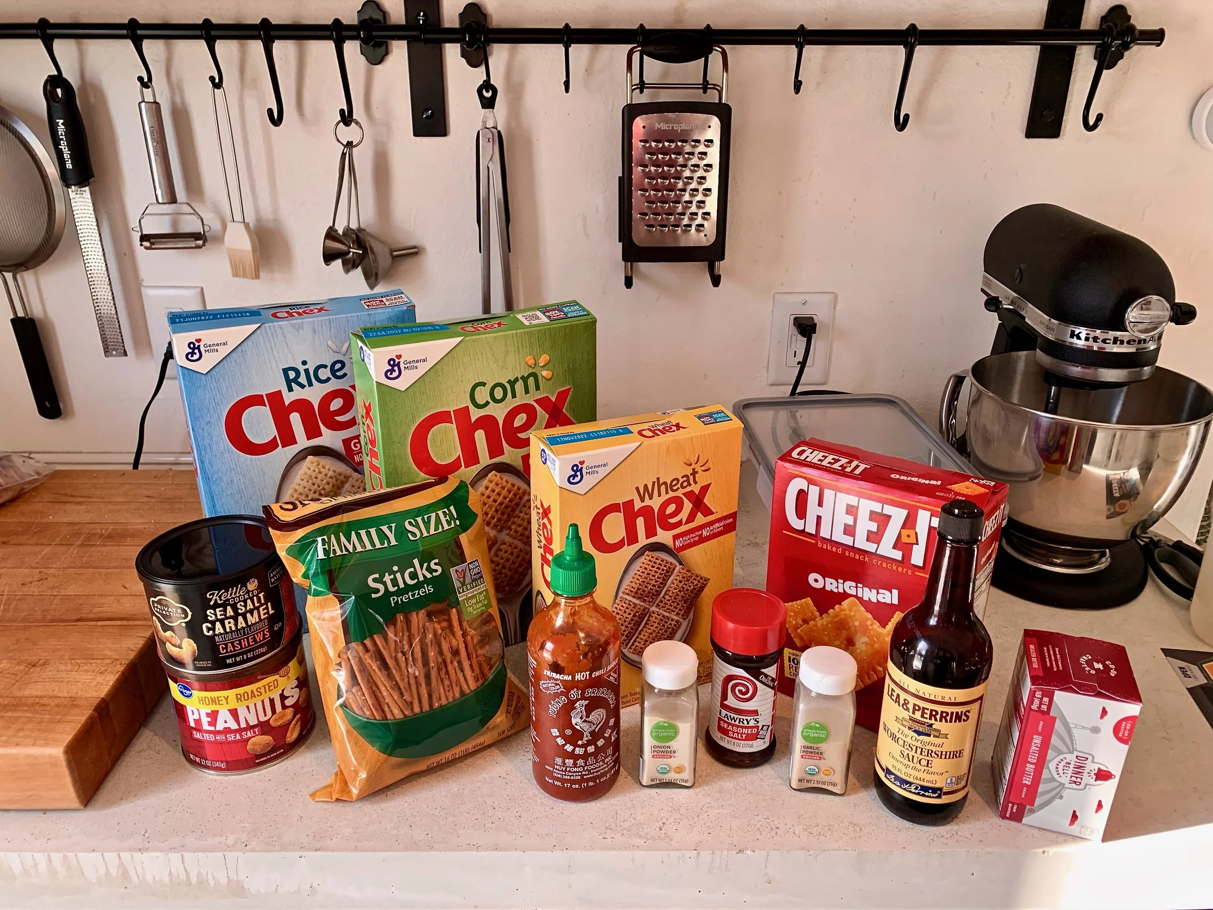 Measured ingredients for Chex party mix on a kitchen counter including Rice Chex, Corn Chex, Wheat Chex, pretzels, mixed nuts, bagel chips, butter, Worcestershire sauce, and spices in small bowls
