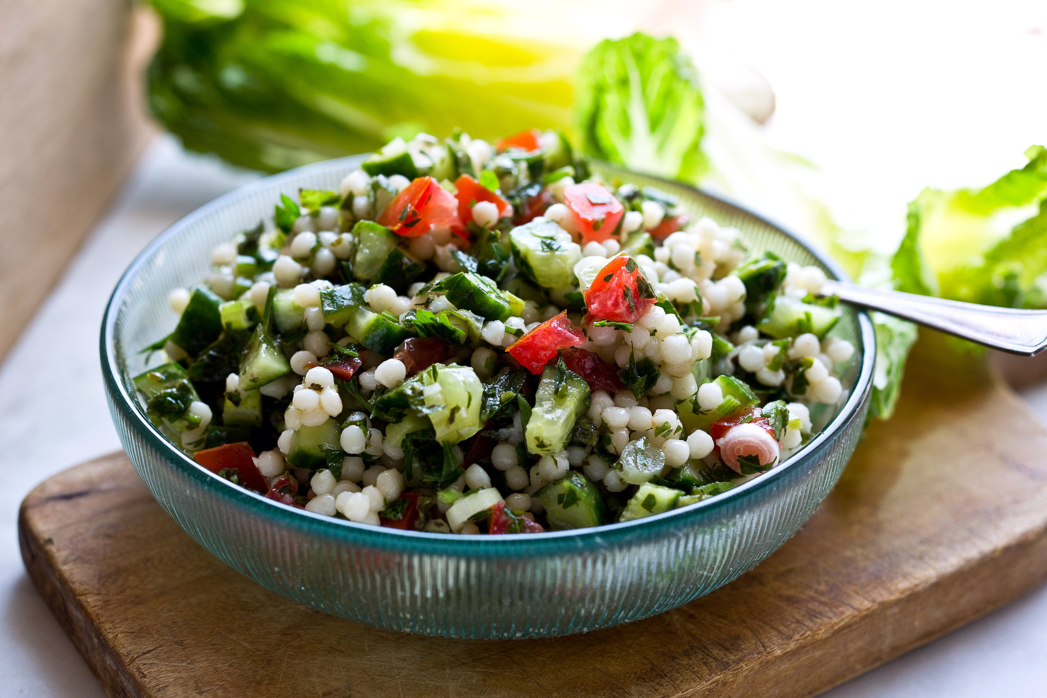 Measured ingredients for Israeli couscous salad on a kitchen counter, including pearl couscous, cucumbers, cherry tomatoes, chickpeas, herbs, feta, lemon, tahini, and olive oil