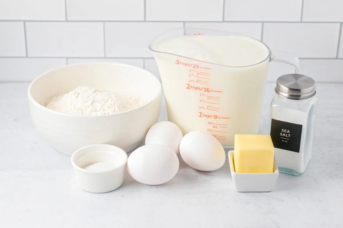 Measured ingredients for Swedish pancakes on a stone kitchen counter including flour, eggs, milk, sugar, vanilla, salt, and melted butter in a small bowl