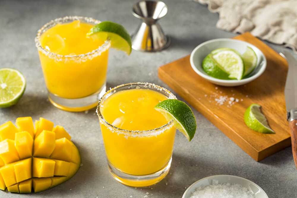 Measured ingredients for a mango margarita including mango chunks, tequila, fresh limes, orange liqueur, agave nectar, and a small dish of chili-lime salt on a bright kitchen counter