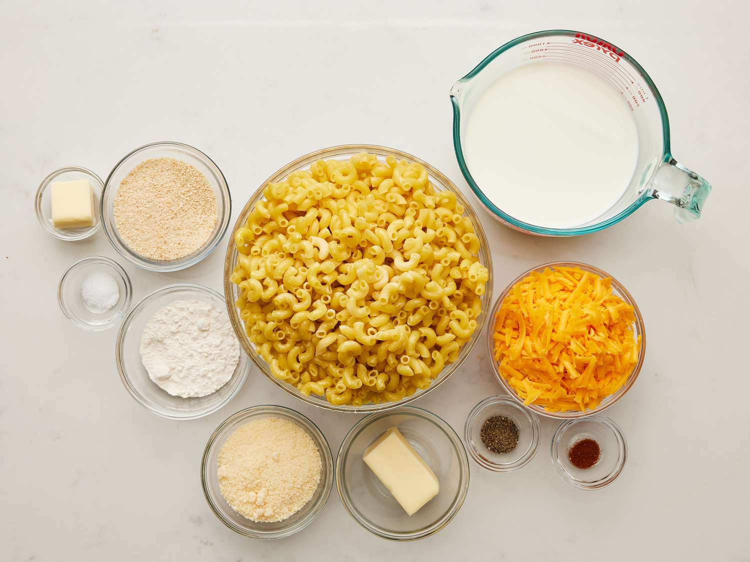 Measured ingredients for baked macaroni and cheese arranged on a tidy kitchen counter, including shredded cheddar, milk, pasta, butter, flour, and breadcrumbs