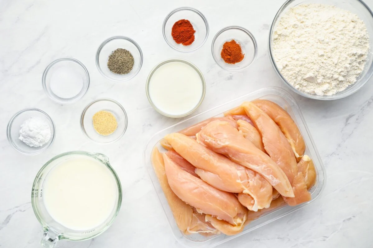 Measured ingredients for buttermilk chicken tenders including buttermilk, chicken tenders, flour, cornstarch, spices, and hot sauce arranged on a bright kitchen counter