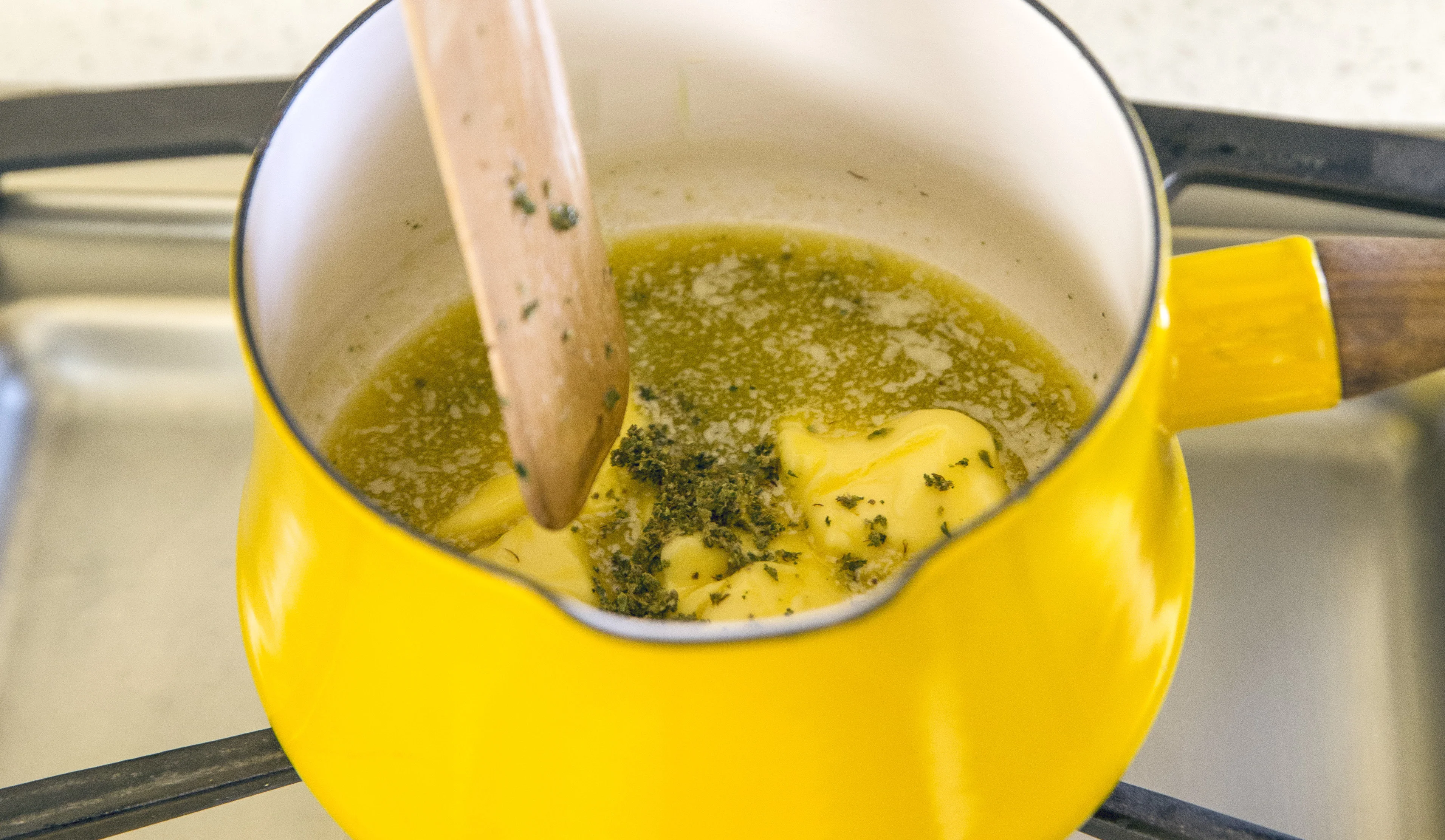 Measured ingredients for cannabutter on a kitchen counter including unsalted butter, dried cannabis, water, and a cheesecloth-lined strainer, bright home kitchen lighting