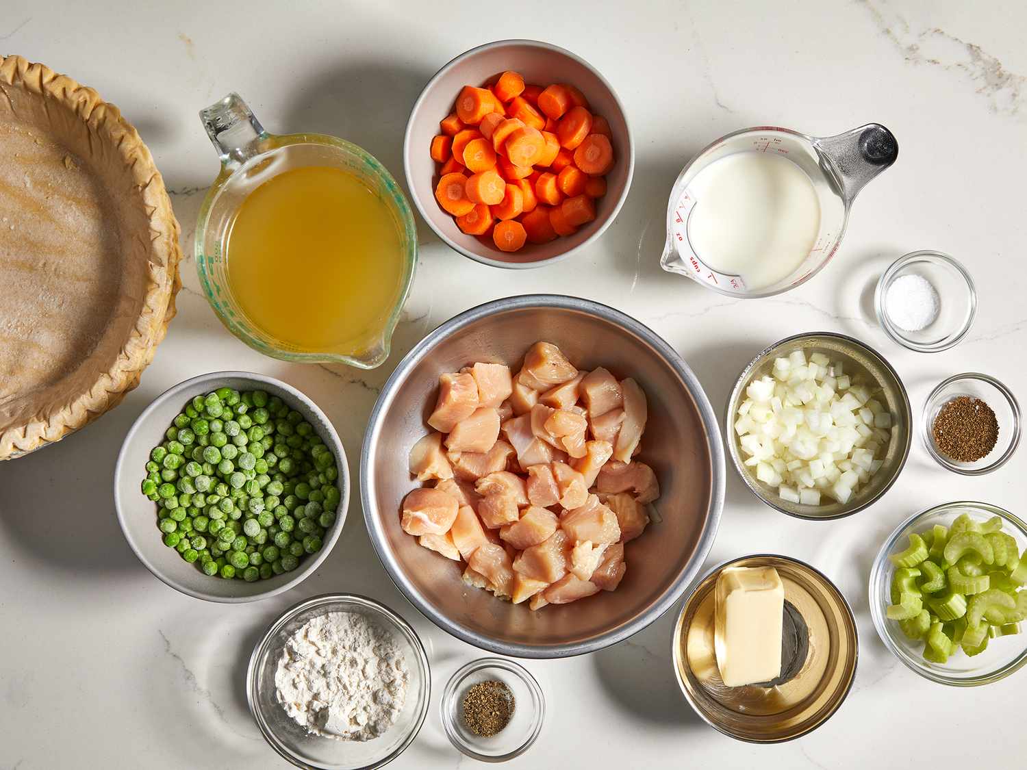 Measured ingredients for chicken pot pie on a bright kitchen counter, including shredded chicken, carrots, peas, celery, onion, chicken broth, milk, and a pie crust