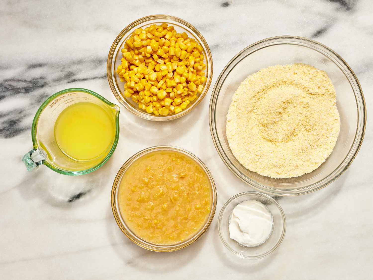 Measured ingredients for cornbread casserole on a kitchen counter including cornbread mix, eggs, sour cream, canned corn, butter, and shredded cheese