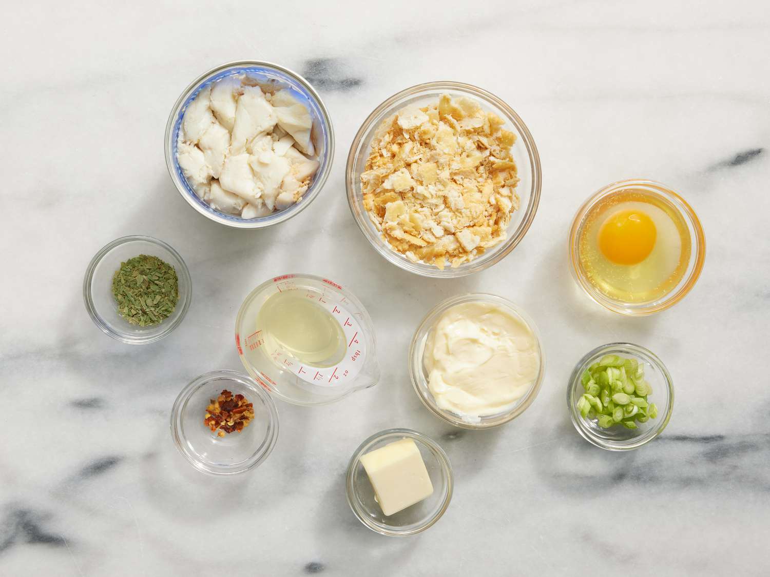 Measured ingredients for crab cakes on a bright kitchen counter including jumbo lump crab, mayonnaise, mustard, Old Bay seasoning, eggs, parsley, lemon, and panko in small bowls