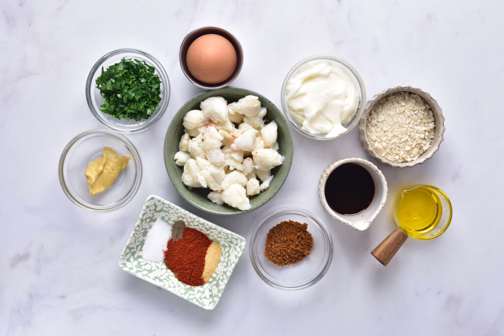 Measured ingredients for crab cakes on a kitchen counter including lump crab meat, crackers, egg, mayonnaise, Dijon mustard, lemon, and herbs in small bowls