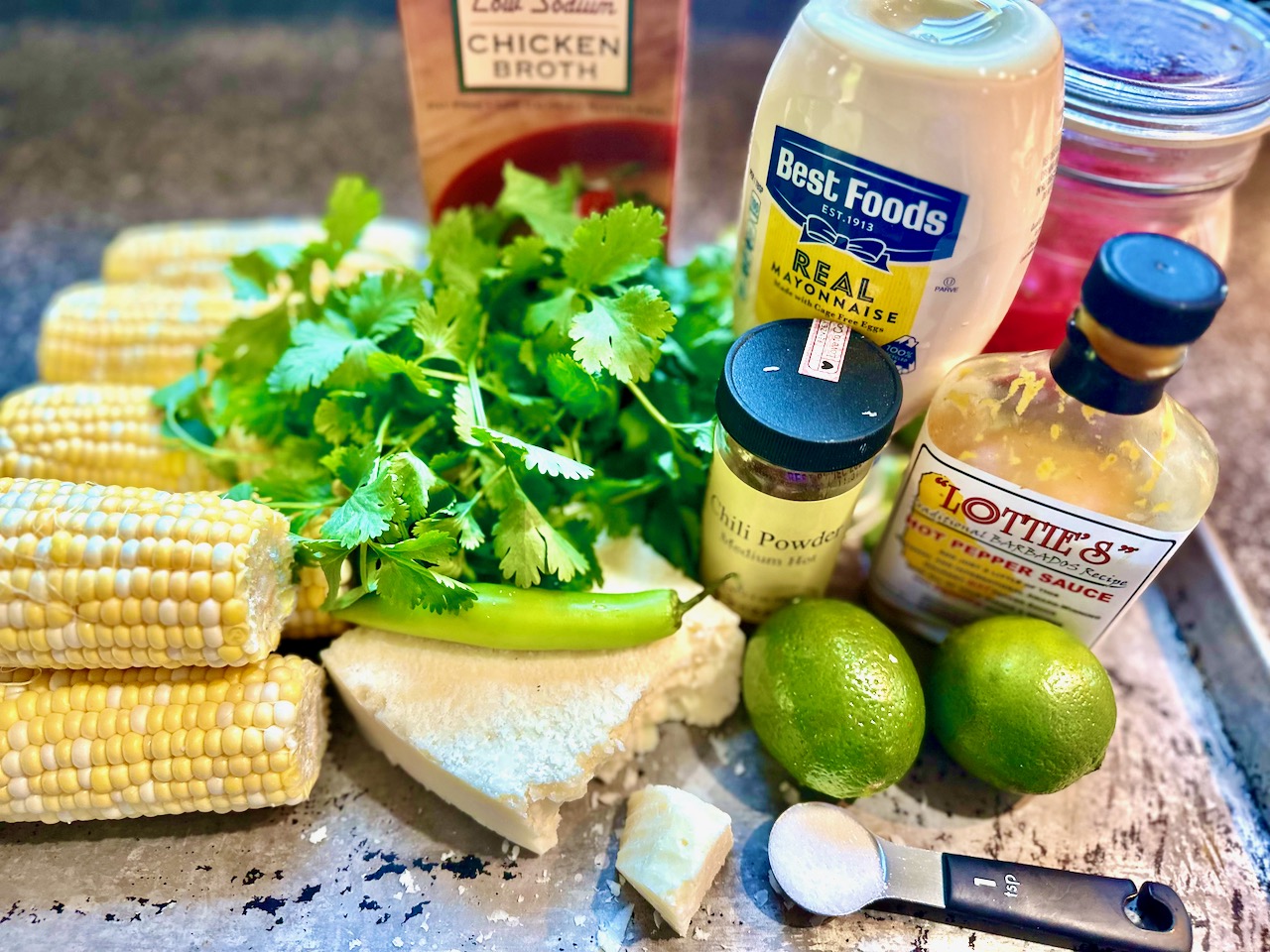 Measured ingredients for elote dip including corn kernels, sour cream, mayonnaise, lime, cotija cheese, cilantro, jalapeno, and spices arranged on a clean kitchen counter
