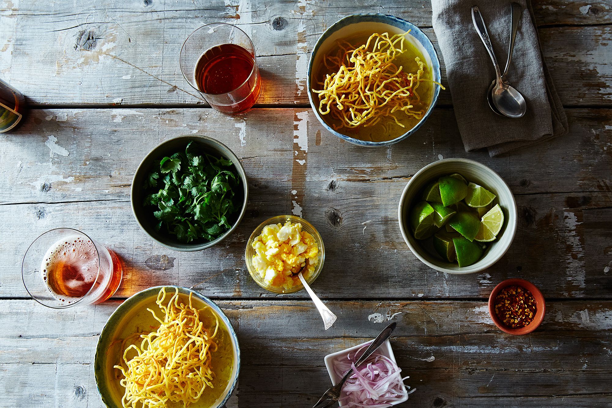 Measured ingredients for khao soi arranged on a kitchen counter including coconut milk, curry paste, chicken thighs, egg noodles, chicken broth, aromatics, lime, and garnishes