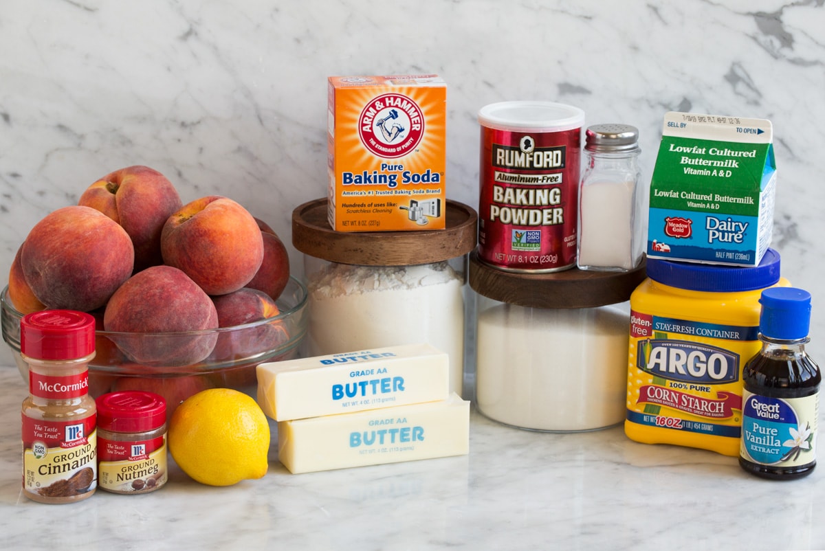 Measured ingredients for peach cobbler including sliced peaches, flour, sugar, brown sugar, butter, cinnamon, and buttermilk on a bright kitchen counter