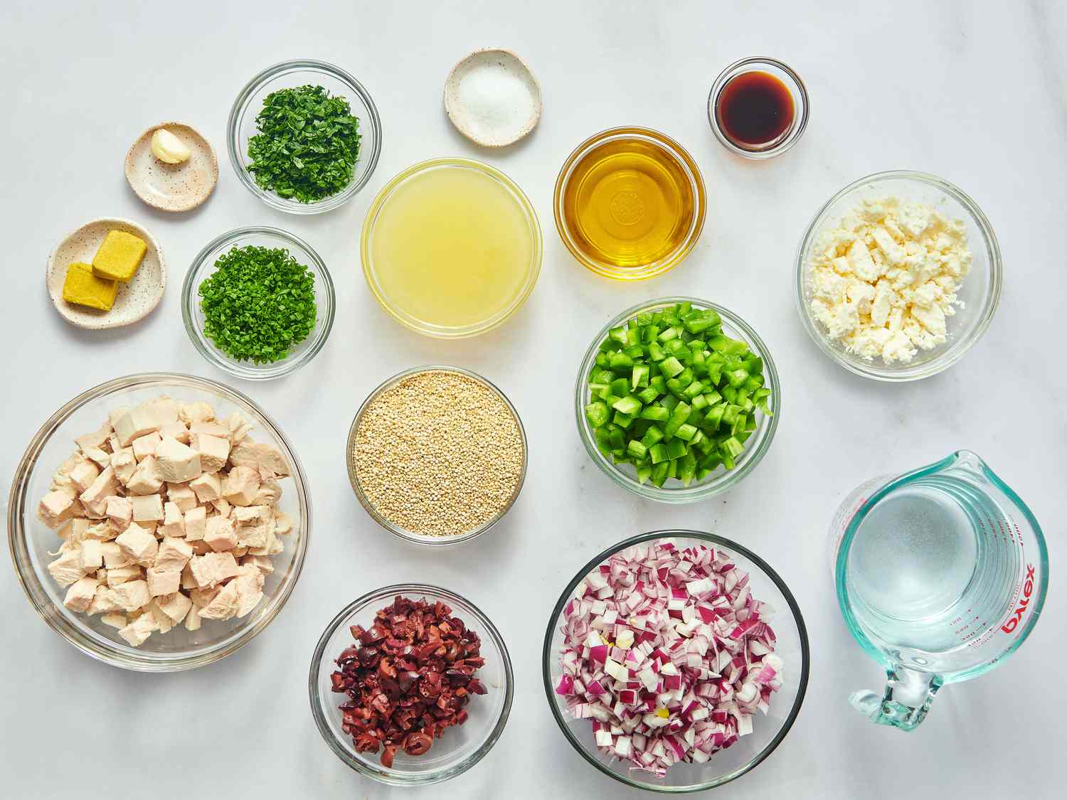 Measured ingredients for quinoa salad on a kitchen counter including cooked quinoa, chickpeas, diced cucumber, chopped herbs, pistachios, feta, lemon, and olive oil in small bowls