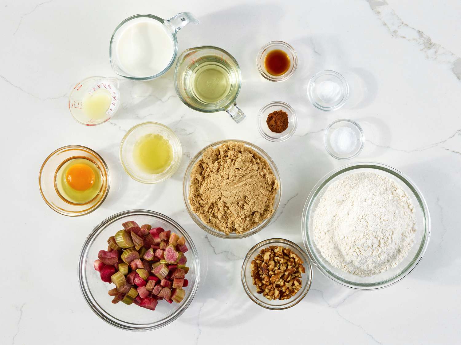 Measured ingredients for rhubarb bread on a kitchen counter including chopped rhubarb in a bowl, flour, sugar, eggs, yogurt, oil, and spices arranged in small dishes