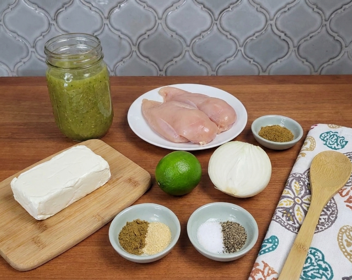 Measured ingredients for salsa verde chicken on a kitchen counter including salsa verde jar, chicken breasts, cream cheese, lime, onion, and spices