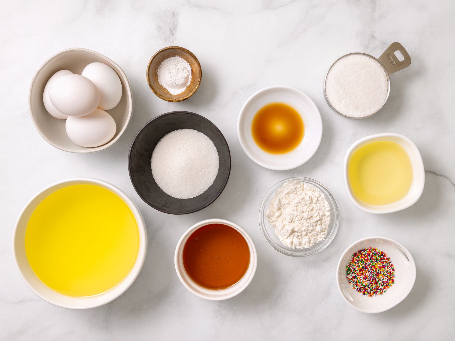 Measured ingredients for struffoli on a wooden kitchen counter, including flour, eggs, sugar, butter, lemon and orange, honey, and small bowls of sprinkles