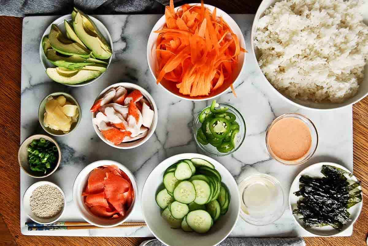 Measured ingredients for sushi bowls including rice, imitation crab, cucumber, avocado, sriracha, mayonnaise, soy sauce, and sesame seeds laid out on a bright kitchen counter