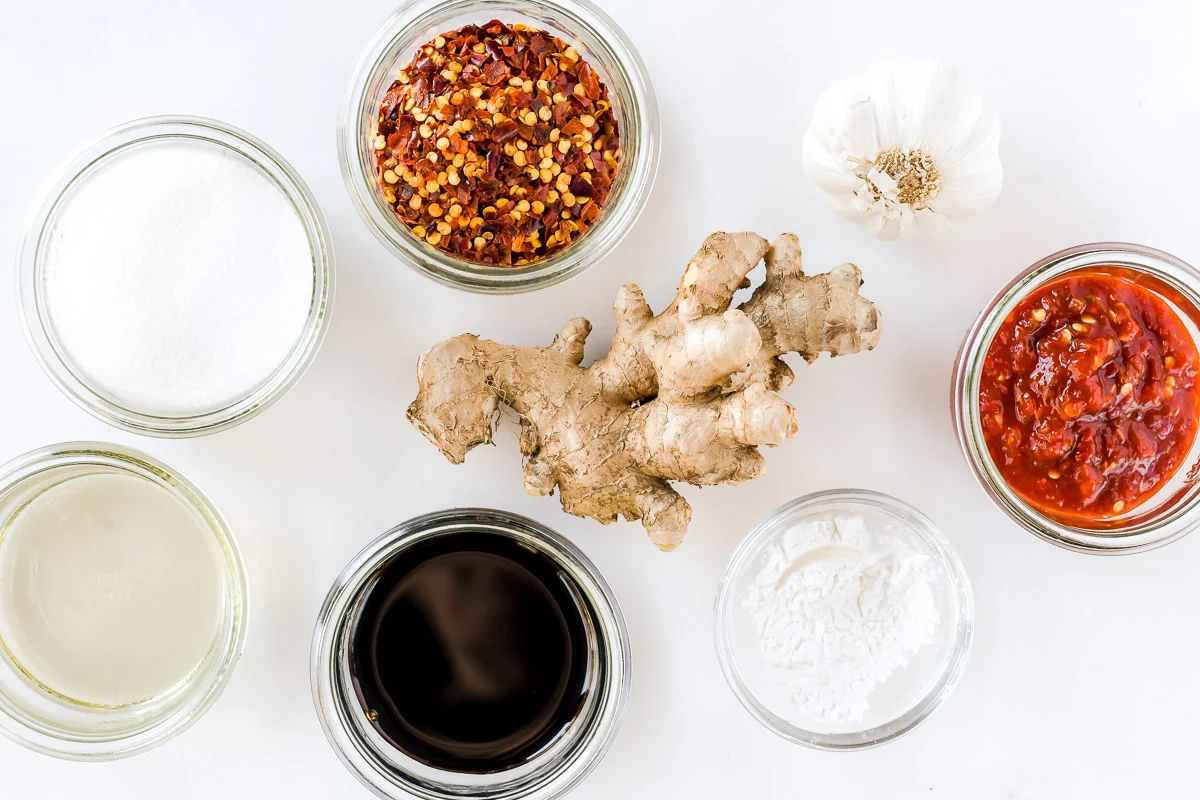 Measured ingredients for sweet chili sauce on a kitchen counter, including rice vinegar, sugar, garlic, red pepper flakes, and cornstarch in small bowls
