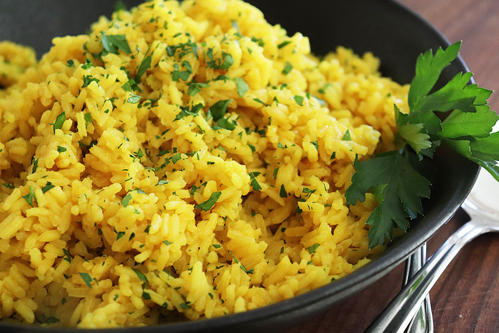Measured ingredients for turmeric rice on a light kitchen counter including basmati rice, ground turmeric, garlic, butter, broth, salt, pepper, lemon, and parsley