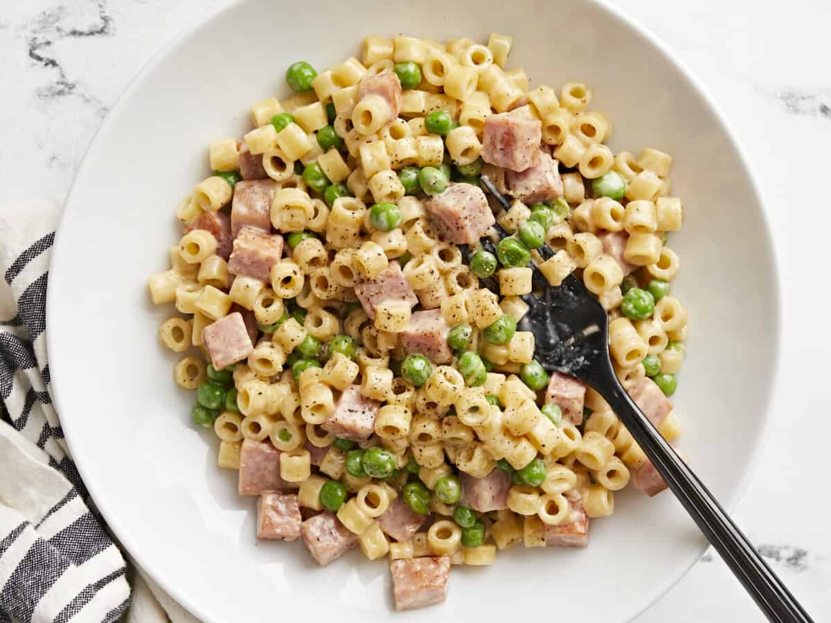 Measured ingredients on a home kitchen counter including pasta, diced ham, frozen peas, garlic, cream, grated Parmesan, and a lemon beside a pot and skillet
